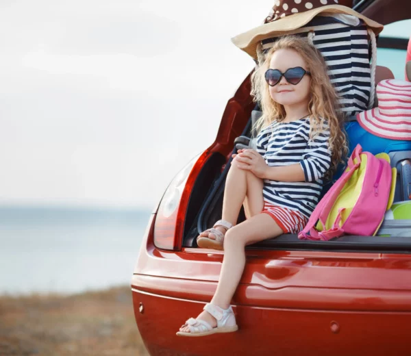 Ein Mädchen sitzt vor Urlaubsgepäck im geöffneten Kofferraum eines Autos: Bei der Reiseapotheke für Kinder gibt es ein paar Punkte zu beachten