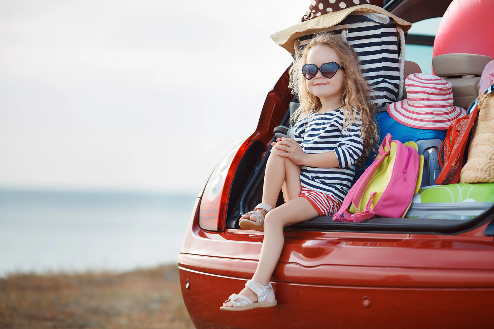 Ein Mädchen sitzt vor Urlaubsgepäck im geöffneten Kofferraum eines Autos: Bei der Reiseapotheke für Kinder gibt es ein paar Punkte zu beachten