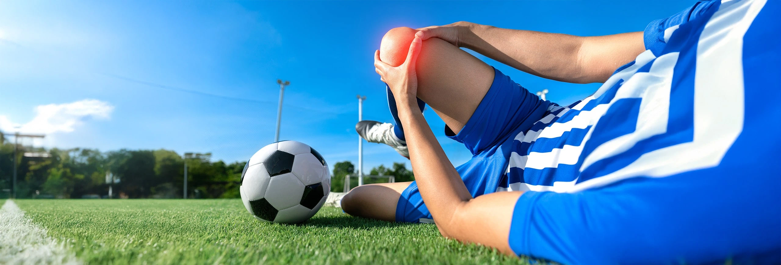 Fußballer Schmerz Knie-min Fußballer liegt am Boden und hält sich schmerzendes Knie. Das Knie ist rot beleuchtet. Neben dem Fußballer liegt ein Fußball.